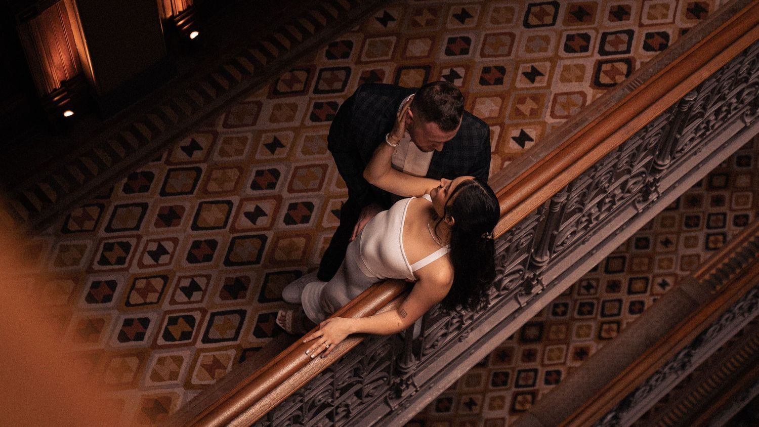 A couple seen from above on an ornate landing, her in white, him in a dark jacket.