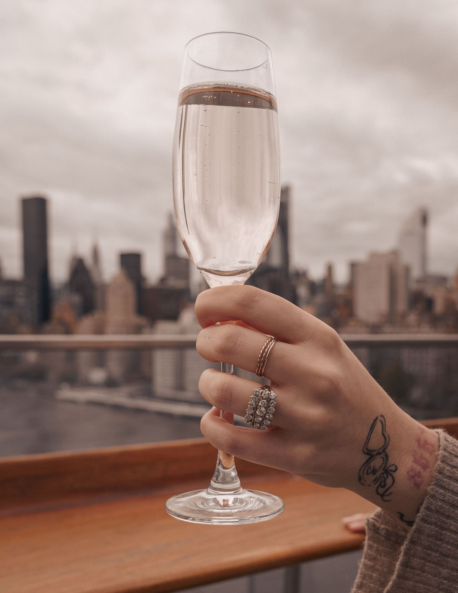 A hand wearing a diamond ring, holding a champagne glass, with the skyline behind it.
